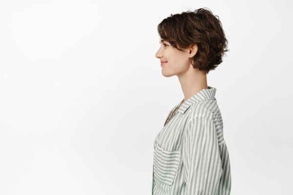 Retrato de perfil de una hermosa mujer sonriente con peinado corto de duendecillo mirando hacia la izquierda con una sonrisa feliz, expresión facial amigable e informal, de pie con una camisa informal sobre fondo blanco.