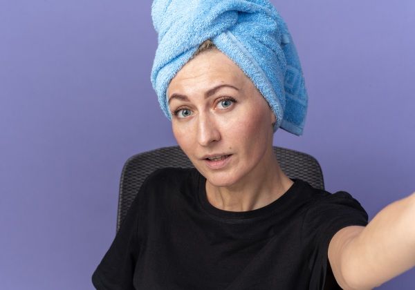 Menina linda e satisfeita sentada à mesa com ferramentas de maquiagem, enxugando o cabelo na toalha, segurando a câmera isolada na parede azul