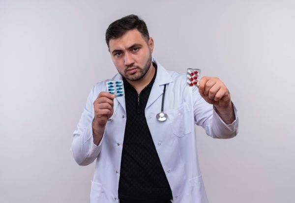Jovem médico barbudo vestindo jaleco branco com estetoscópio segurando bolhas com comprimidos e mostrando para a câmera com uma cara séria