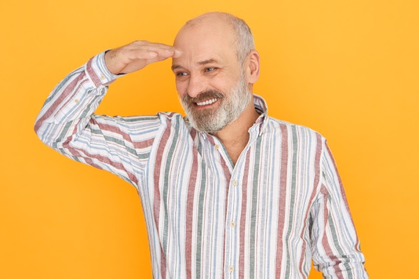 transplante capilar aos 50 - Homem idoso atraente e alegre com barba grisalha e careca posando isolado, mantendo a mão sobre os olhos para se proteger