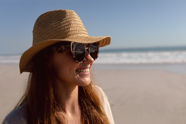 Cuidar do cabelo no verão - mulher na praia protegendo o cabelo do sol com um chapéu